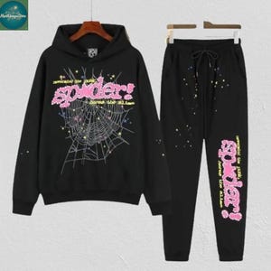 May include: Black hoodie and sweatpants set. The hoodie features a pink "Spider!" graphic with a spiderweb design. The sweatpants have a matching pink graphic on the leg and a drawstring waist. The set is made of a soft material.