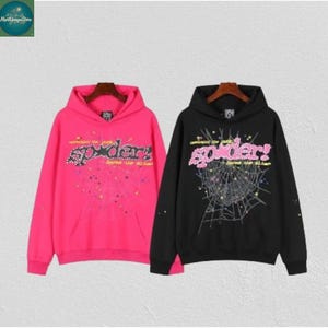 May include: Two hooded sweatshirts, one hot pink and one black, each featuring the word "spider" in a stylized font with a spiderweb design. The pink sweatshirt has a matching pink hood and a front pocket. The black sweatshirt has a matching black hood and a front pocket.