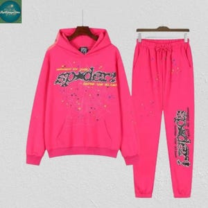 May include: A bright pink hoodie and matching sweatpants set. The hoodie features a front pocket and the word "spider" in a stylized font with rhinestone accents. The sweatpants have a drawstring waist and the word "spider" on the leg.