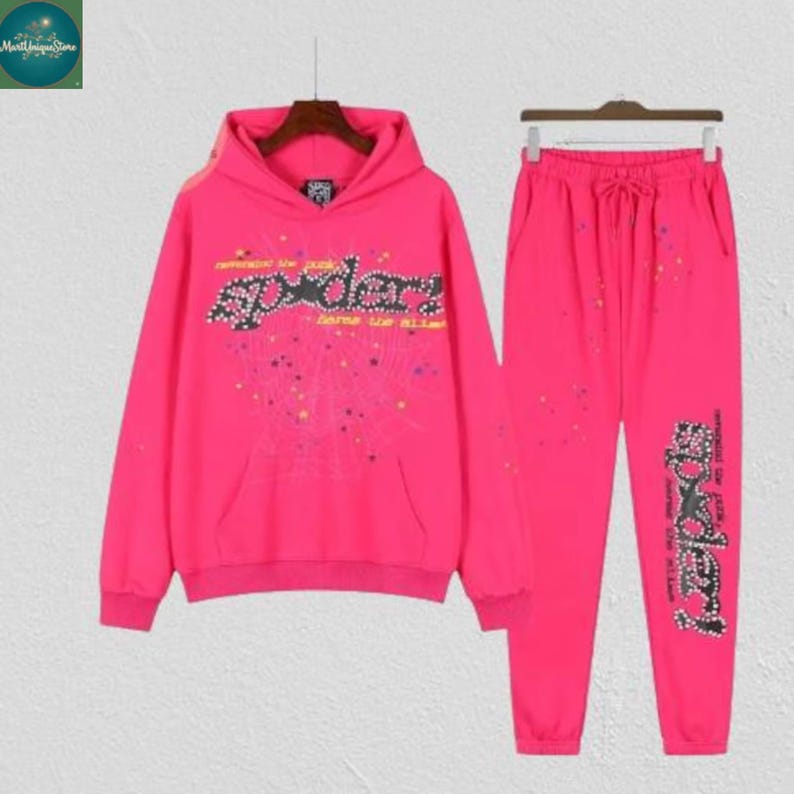 May include: A bright pink hoodie and matching sweatpants set. The hoodie features a front pocket and the word "spider" in a stylized font with rhinestone accents. The sweatpants have a drawstring waist and the word "spider" on the leg.