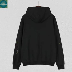 May include: A black hoodie with a hood and long sleeves. The sleeves have a constellation design in yellow. The hoodie is hanging on a hanger against a light-colored wall. The hoodie is a casual garment.