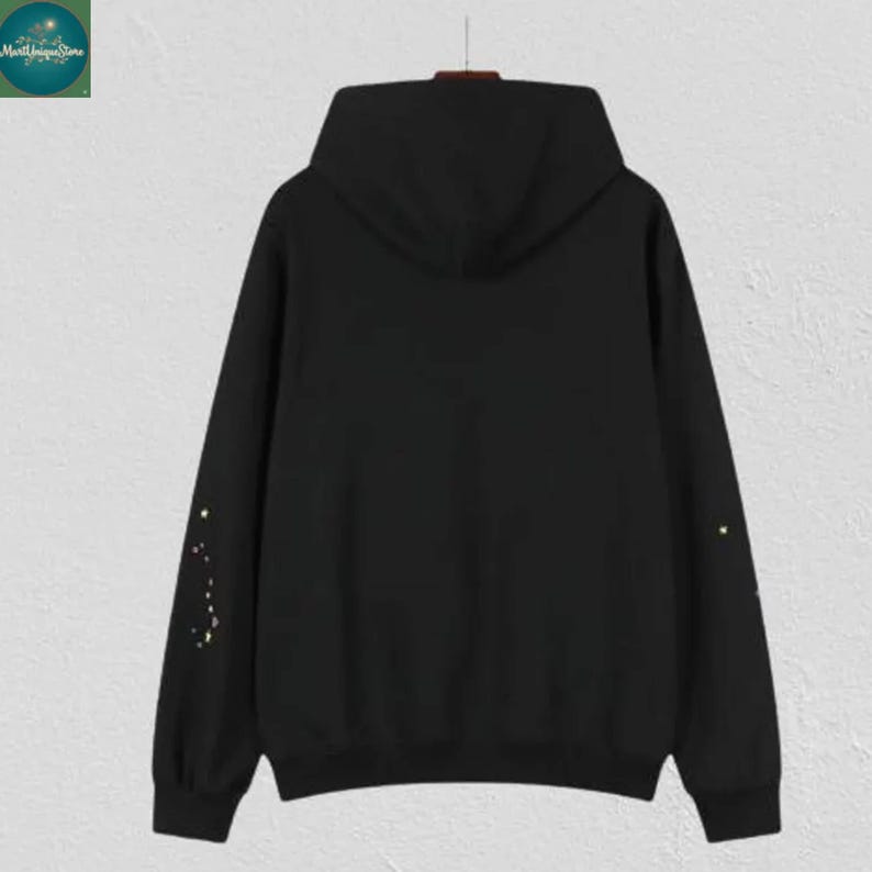 May include: A black hoodie with a hood and long sleeves. The sleeves have a constellation design in yellow. The hoodie is hanging on a hanger against a light-colored wall. The hoodie is a casual garment.