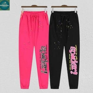 May include: Two pairs of sweatpants, one hot pink and one black, hanging on a rack. Both have a drawstring waist, pockets, and a decorative design with the word "Spidey!" in a stylized font. The pink pants have colorful splatter details.