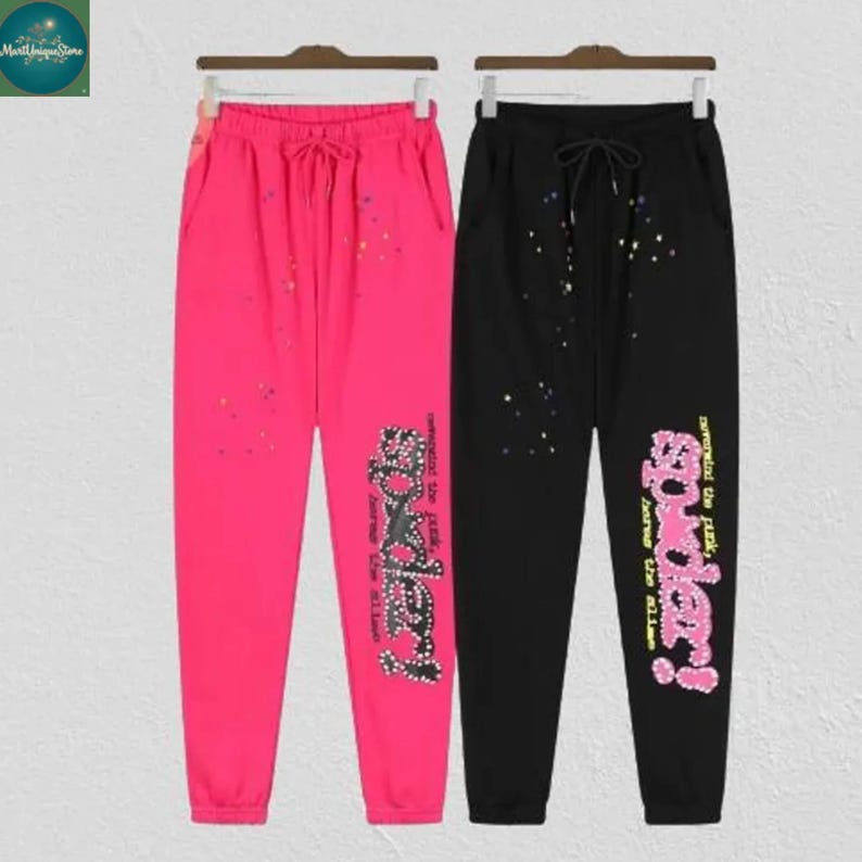 May include: Two pairs of sweatpants, one hot pink and one black, hanging on a rack. Both have a drawstring waist, pockets, and a decorative design with the word "Spidey!" in a stylized font. The pink pants have colorful splatter details.