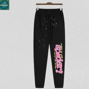 May include: Black sweatpants with a drawstring waist and colorful splatter paint design. The pants feature the text "Spider!" in pink and yellow, with additional text in yellow. The sweatpants are hanging on a wooden hanger.
