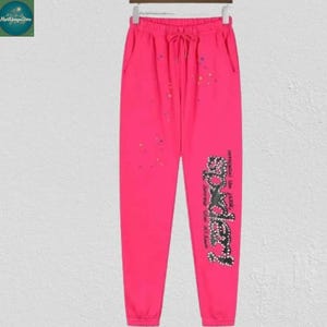 May include: Bright pink sweatpants with a drawstring waist and elastic cuffs. The pants feature a black and white graphic on the left leg that says "bodley" and "remember the faith, journey". Small colorful dots are scattered across the front.