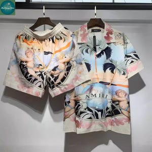 May include: A two-piece matching set featuring a short-sleeved button-down shirt and shorts. The set has a cream background with a colorful print of cherubs, palm trees, and a globe. The shirt has the brand name "AMIRI" printed on it.