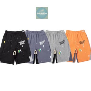 Puede incluir: Cuatro pares de pantalones cortos en negro, azul grisáceo, gris y naranja. Cada par presenta detalles de salpicaduras de pintura y el texto "GALLERY DEPT.". Los pantalones cortos tienen cintura con cordón y diseño de abertura.