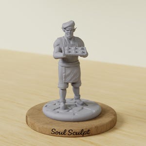 May include: A grey, unpainted miniature figurine of a baker with pointed ears, wearing a chef hat and apron, holding a rectangular tray of six small round pastries. The figure stands on a circular stone-textured base with the text Soul Sculpt.