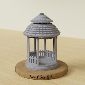 Puede incluir: Un cenador en miniatura gris con un techo escalonado y una barandilla de valla. El cenador está sobre una base de madera redonda. El texto "Soul Sculpt" está impreso en la base.