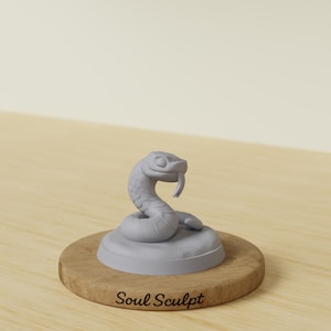 Könnte beinhalten: Eine hellgraue Schlangenfigur mit gewundenem Körper und herausgestreckter Zunge, montiert auf einem runden Sockel. Der Sockel steht auf einer runden Holzplatte, auf deren Vorderseite der Text Soul Sculpt in einer dunklen Schreibschrift eingraviert ist.