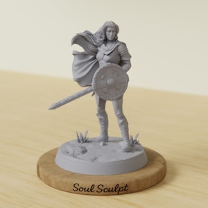 以下が含まれることがあります： 剣と盾を持った女性戦士の灰色のフィギュアが、円形の台座の上に立っています。フィギュアは鎧とマントを着ています。台座は木製のスタンドに置かれています。「Soul Sculpt」の文字が台座に印刷されています。