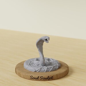 Op de afbeelding: Een grijze cobra-sculptuur met een open mond, klaar om toe te slaan. De slang is gemonteerd op een ronde, houten basis met de tekst "Soul Sculpt" in een cursief lettertype. De sculptuur is gedetailleerd en realistisch.