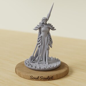 May include: A gray figurine of a woman in a flowing dress, holding a spear and a staff with a decorative head. The figurine is on a wooden base with the text "Soul Sculpt". The figure has feather-like details on the shoulders and dress.