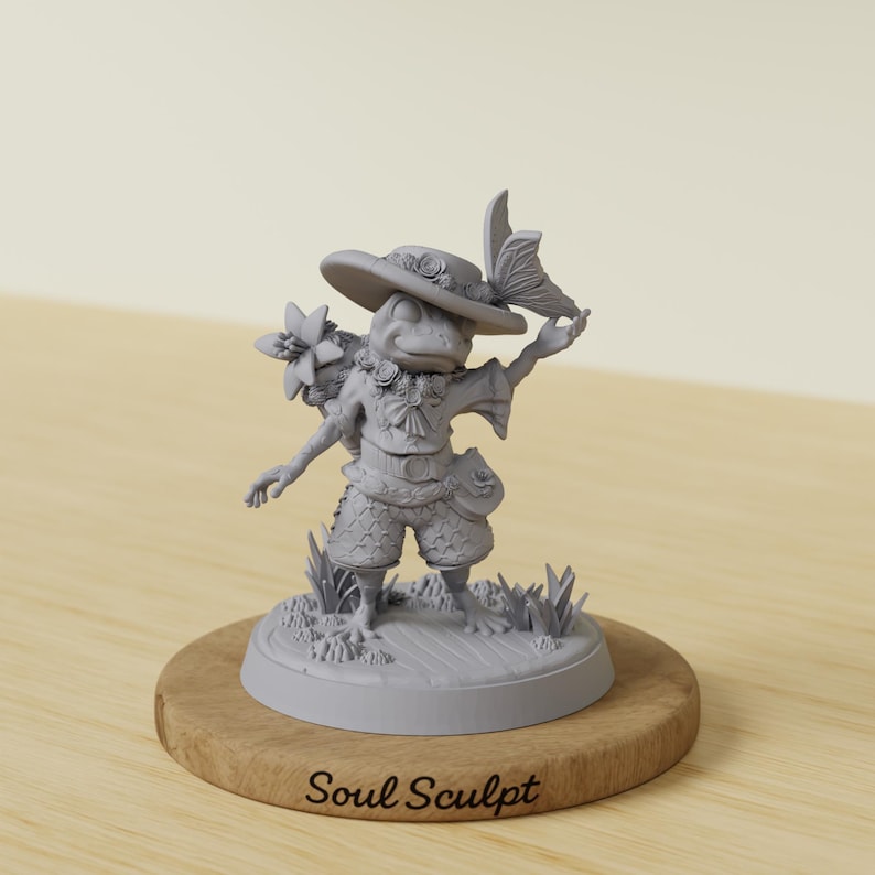 Op de afbeelding: Een grijze kikkerfiguur met een hoed, korte broek en een bloemstuk. De kikker houdt een vlinder vast en staat op een houten voet met de tekst "Soul Sculpt". Het beeldje is gedetailleerd en grillig.