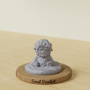 May include: A grey sculpture of a seated creature with a textured head and smiling face. The sculpture sits on a round wooden base with the text "Soul Sculpt" in a cursive font. The background is a light beige.