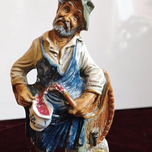 Puede incluir: Figura de cerámica de un hombre vestido de viticultor, vertiendo líquido rojo de una jarra pequeña. La figura tiene un tapón de corcho en la parte superior y está pintada en tonos marrones, azules y blancos. La base está diseñada para parecer un ladrillo.