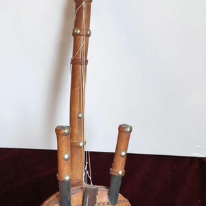 Könnte beinhalten: Handgefertigtes Saiteninstrument mit lederbezogenem Korpus und Holzhals. Das Instrument hat dekorative Metallnieten und eine geflochtene Lederschlaufe. Der Korpus weist ein kompliziertes Muster auf.