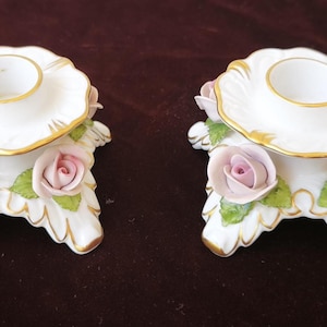 Puede incluir: Par de candelabros de porcelana blanca con detalles dorados y rosas rosas. Cada uno presenta una parte superior festoneada y una base adornada con rosas y hojas verdes. Un artículo decorativo.