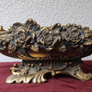 May include: An antique-style, gold-coloured decorative bowl with a floral design. The bowl features three large rose blossoms and intricate leaf details. It has handles and sits on a decorative base.