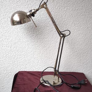 Könnte beinhalten: Eine verchromte Schreibtischlampe mit verstellbarem Arm und rundem Sockel. Die Lampe hat einen gewölbten Schirm und ein schwarzes Netzkabel. Die Lampe steht auf einer weinroten Oberfläche.