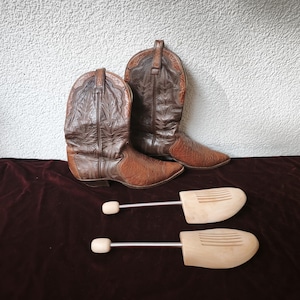 Könnte beinhalten: Ein Paar braune Leder-Cowboystiefel mit Ziernähten am Schaft und strukturierten Vorderkappen, begleitet von zwei hellen Holz-Schuhspannern mit Metallfedern, die dazu dienen, die Form des Schuhwerks beizubehalten.