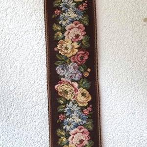 May include: A long, rectangular tapestry with a dark brown background and a floral pattern. The flowers are in shades of pink, yellow, and blue. The tapestry is approximately 76 cm long and 15 cm wide, with a decorative metal top and a gold tassel.