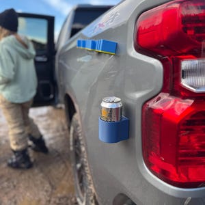 Könnte beinhalten: Ein blauer magnetischer Getränkedosenhalter an der Seite eines grauen Lastwagens befestigt. Eine silberne Dose befindet sich im Halter. Ein blauer und gelber Magnetstreifen ist ebenfalls am LKW befestigt. Der Hintergrund zeigt eine Person und einen bewölkten Himmel.