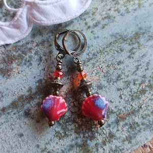 Könnte beinhalten: Ohrringe mit roten und blauen muschelförmigen Keramikperlen. Die Ohrringe haben bronzefarbene Metallakzente, darunter dekorative Perlen und einen Klappverschluss. Kleine orangefarbene und rote Perlen.