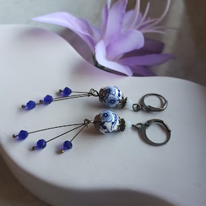 Könnte beinhalten: Ohrringe mit weißen und blauen Keramikperlen mit Blumenmuster. Jeder Ohrring hat drei dunkelblaue Kristallperlen und antike bronzefarbene Metallakzente und Ohrhaken. Im Hintergrund befindet sich eine lila Blume. Ein dekoratives Accessoire.