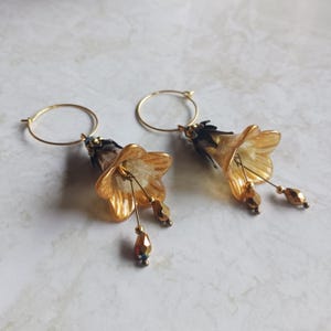 Könnte beinhalten: Ein Paar goldfarbene Creolen mit hängenden blumenförmigen Anhängern. Die durchscheinenden bernsteinfarbenen Blütenblätter sind mit dunklen Metallkappen und kleinen, facettierten Goldperlen verziert, die an dünnen Drähten aus der Mitte hängen.
