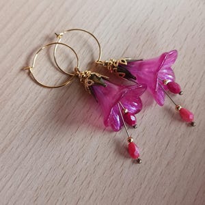 Könnte beinhalten: Ein Paar handgefertigte Ohrringe mit fuchsiafarbenen Blumenanhängern mit goldfarbenen Akzenten und Creolenverschlüssen. Die Blumenanhänger haben ein durchscheinendes Aussehen und sind mit hängenden roten Perlen verziert. Die Ohrringe liegen auf einer hellen Holzoberfläche.