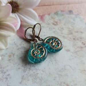 Könnte beinhalten: Türkisfarbene Ohrringe mit goldenem Spiralmuster. Die Ohrringe haben einen Klappverschluss und sind vor einem weichen, floralen Hintergrund platziert. Die Ohrringe sind etwa 2,5 cm lang.
