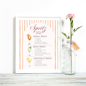 K&ouml;nnte beinhalten: Ein gerahmter Druck mit einem pfirsichfarbenen und wei&szlig;en gestreiften Hintergrund zeigt Cocktailrezepte, darunter Aperol, Hugo und Limoncello Spritz. Der Text "Spritz Bar" steht oben. Rechts ist eine klare Glasflasche mit einer rosa Rose.
