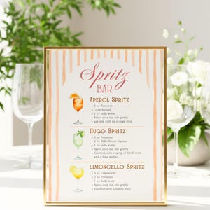 Puede incluir: Un letrero enmarcado en oro muestra un menú de "Spritz Bar". El menú presenta recetas de cócteles Aperol, Hugo y Limoncello Spritz, con ilustraciones de acuarela de cada bebida. El fondo tiene rayas verticales melocotón y blancas.