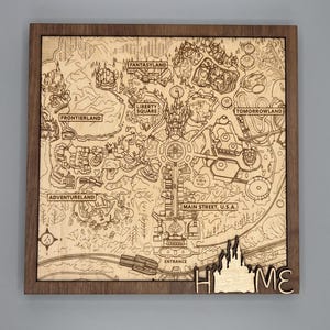 May include: A square wooden wall art piece featuring a laser-engraved map of a theme park. The map includes labels for Frontierland, Fantasyland, Liberty Square, Tomorrowland, Adventureland, and Main Street, U.S.A. The word HOME is at the bottom.