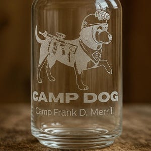 Könnte beinhalten: Klares, becherförmiges Trinkglas mit einem geätzten Design eines Hundes, der einen Helm trägt und ein Gewehr trägt. Der Text "CAMP DOG" und "Camp Frank D. Merrill" sind unter dem Hunde-Bild geätzt.