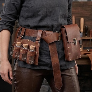 May include: A brown leather belt with multiple pouches and small glass bottles. The belt has a large buckle and a rectangular pouch with a metal clasp. The belt is worn over brown leather pants and a dark gray shirt.