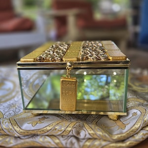 May include: A rectangular glass jewelry box with gold detailing and a gold bar charm. The box has a gold-coloured top with textured detailing. The charm reads "GOLD COPY" and hangs from the front of the box.