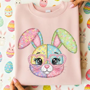 May include: A light pink sweatshirt featuring a colorful patchwork bunny face with heart-shaped glasses. The bunny has large eyes and long ears with floral patterns. The design is centered on the front of the sweatshirt, perfect for Easter.