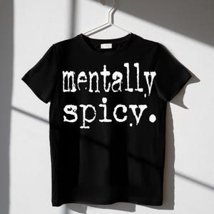 May include: Black t-shirt with the white text "mentally spicy." The shirt is hanging on a white hanger against a white wall. The text is in a bold, slightly distressed font.