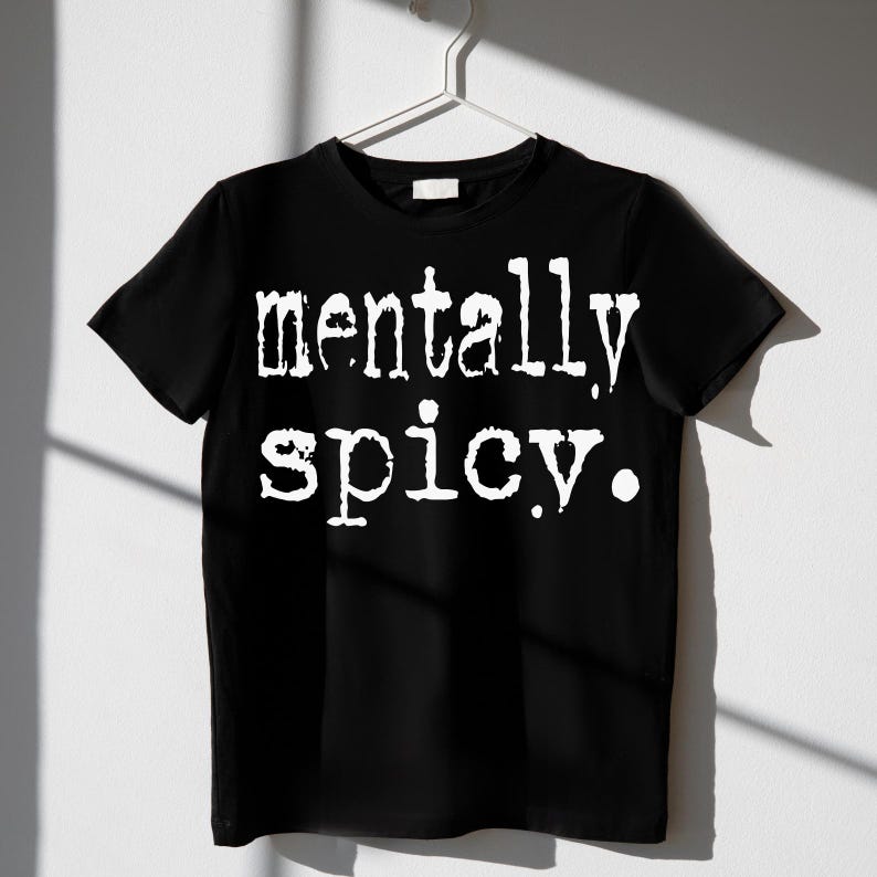 May include: Black t-shirt with the white text "mentally spicy." The shirt is hanging on a white hanger against a white wall. The text is in a bold, slightly distressed font.