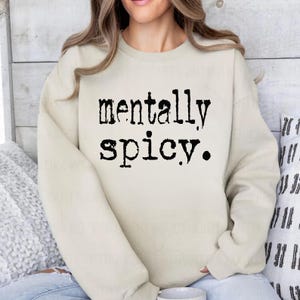 May include: A cream-colored sweatshirt with the words "mentally spicy" printed in black, bold letters. The sweatshirt has a crew neck and long sleeves. The text is centered on the front of the shirt.