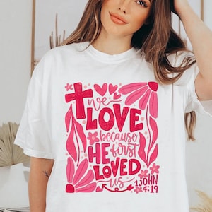 May include: White t-shirt with a pink floral and text design. The text reads "We Love Because He First Loved Us" with the Bible verse 1 John 4:19. The design includes a cross, hearts, and flowers.