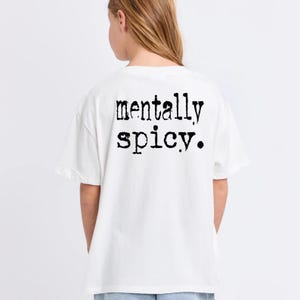 May include: White t-shirt with the phrase "mentally spicy" printed in black, bold letters. The shirt is a casual style, likely made of cotton or a similar fabric. The text is centered on the back of the shirt.