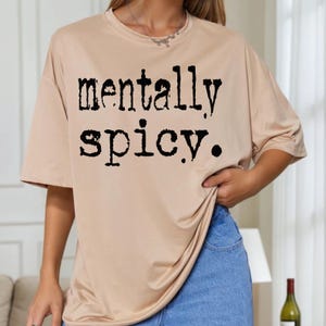 May include: A tan t-shirt with the words "mentally spicy" printed in black, bold letters. The shirt has short sleeves and a relaxed fit. The model is wearing blue jeans.