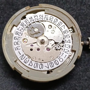 Puede incluir: Un movimiento de reloj mecánico circular con una placa de metal plateado, engranajes, tornillos y rubíes expuestos. Un disco de fecha blanco con números negros impresos del 1 al 31 rodea el mecanismo y tiene una pequeña corona de metal.