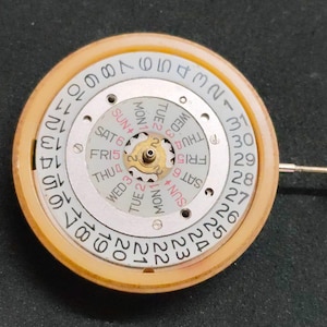 May include: A circular watch movement mechanism featuring a white outer date ring numbered 1 through 31 in black. The inner day-of-the-week wheel displays abbreviations in black and red text, centered around a small brass gear with a metal stem extending outward.