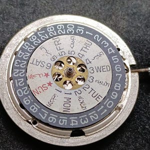 Peut inclure: Un composant de mouvement de montre mécanique circulaire doté d'un disque de date extérieur noir numéroté de 1 à 31 et d'un disque de jour central argenté affichant du texte en anglais et en arabe, avec une tige de remontoir métallique sur le côté.