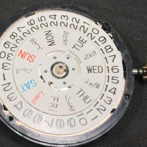 Puede incluir: Un componente de mecanismo de reloj circular con una esfera blanca, números negros del 1 al 31, indicadores de día en inglés y árabe, y una corona metálica en el lado derecho. El mecanismo central es visible, incluyendo el texto 160256S.